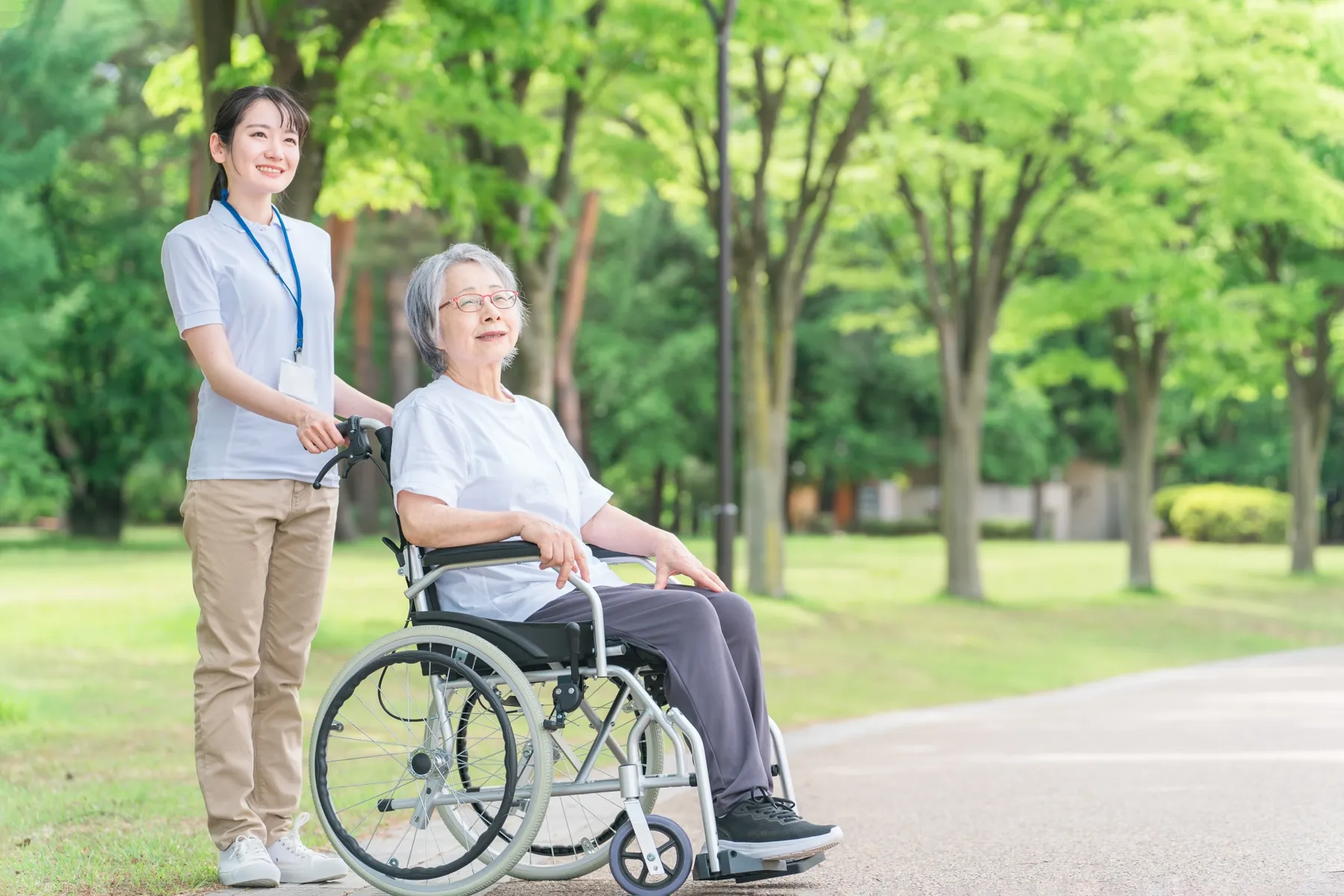新緑の並木道を、車いすの女性に付き添って歩く看護師